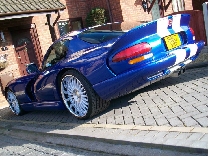 Pistonheads - The image features a sports car that is bright blue with a distinctive checkered pattern running along its flanks. The vehicle is parked on a brick driveway and it appears to have a coupe or convertible design. The car looks sleek and well-maintained, with a curved body that accentuates its speed and performance capabilities. The sun is shining on the car's rear, highlighting its unique color scheme and the shaping of its bodywork.