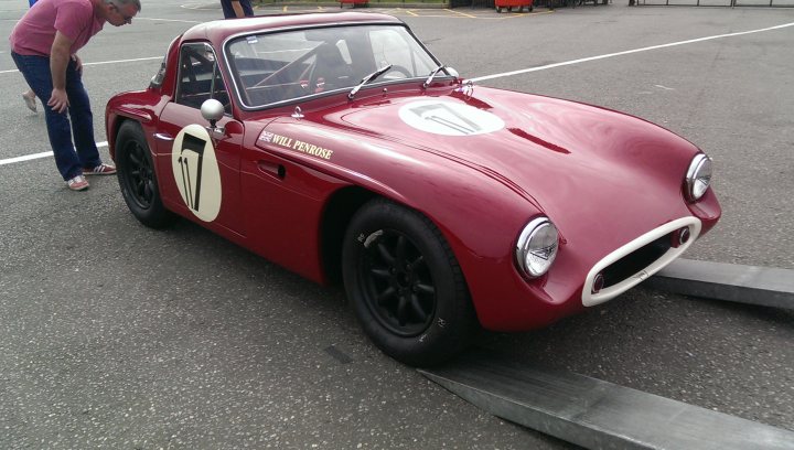 Club race and rally pictures 2016 - Page 2 - UK Club Motorsport - PistonHeads - The image showcases a scene involving a man and a classic red car. The red car, a classic model, is parked on a grey road. The man, who is standing on the pavement, appears to be leaning over the car in an unusual pose. A handrail is visible in front of the car, suggesting that the car might be positioned near an accessible loading or unloading area. In the background, there are several other individuals, but they are too small and distant to make out any specific details about them.