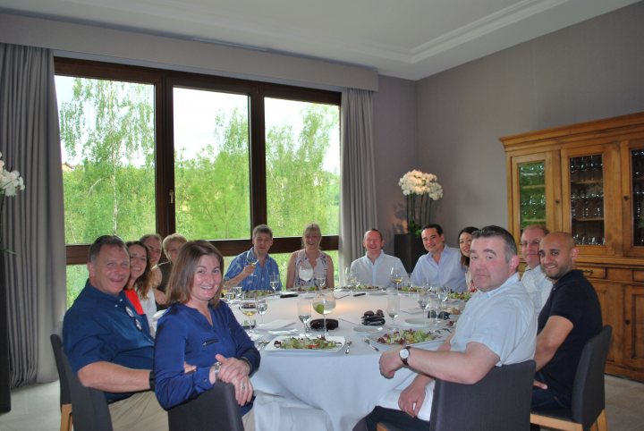 Pistonheads trip to Monaco Historique - Page 2 - Aston Martin - PistonHeads - The image shows a large group of people gathered around a long dining table placed indoors. They are seated on chairs and appear to be engaged in a meal, with the room filled with warmth and conversation. The table is set with various tableware items, including wine glasses, plates, cutlery, and cups. On the table, there are also some food items visible. The atmosphere is friendly and inviting, suggesting a family gathering or a social event. The setting sun outside the large windows gives the room a soft illumination, adding to the intimate and cozy ambiance.