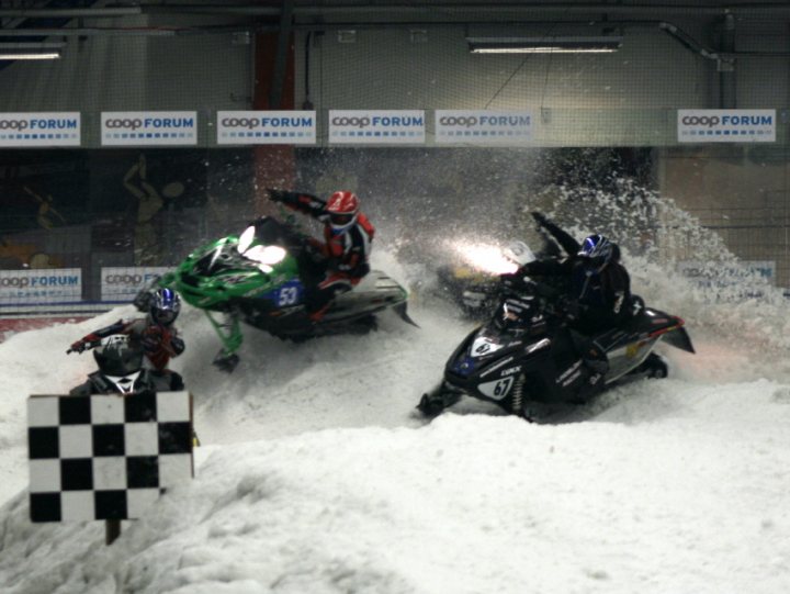 In an indoor setting, three motorcyclists are racing across a snow-covered ground. The snowboarder in front starts to splash into a protective barrier. Their speed and maneuverability are evident as they navigate through the artificial snow. Padded and equipped for the sport, these athletes are engaged in precise and exciting competition.