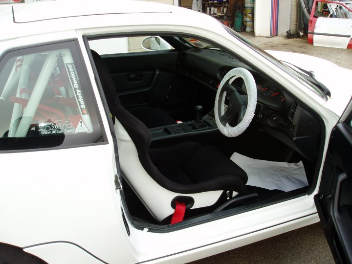 Refreshed Porsche Pistonheads - The image shows the interior of a two-door white car with the driver's side door left open. Inside, the vehicle is equipped with a stereo system, the dashboard features a few controls and dials indicative of a performance car, and there is a driving seat with black upholstery and a logo on the backrest. The car is parked on an asphalt surface, and the rear view is visible in the top left corner of the image. There are some objects inside a garage or workshop in the background, suggesting the vehicle may be undergoing servicing or modification.