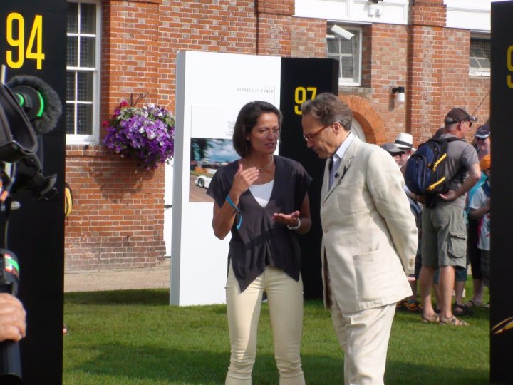 Goodwood FOS on ITV4 - Page 1 - Goodwood Events - PistonHeads - The image captures an outdoor event in a yard in front of a brick building. Two individuals, a man and a woman, are standing in the foreground, engaged in a conversation. The man is dressed in a formal light-colored suit, while the woman sports a casual outfit with a black top. They appear to be friends, given their close proximity and face-to-face engagement. Behind the two individuals, several others are visible, likely attendees of the event, indicating a social gathering or celebration of some sort.