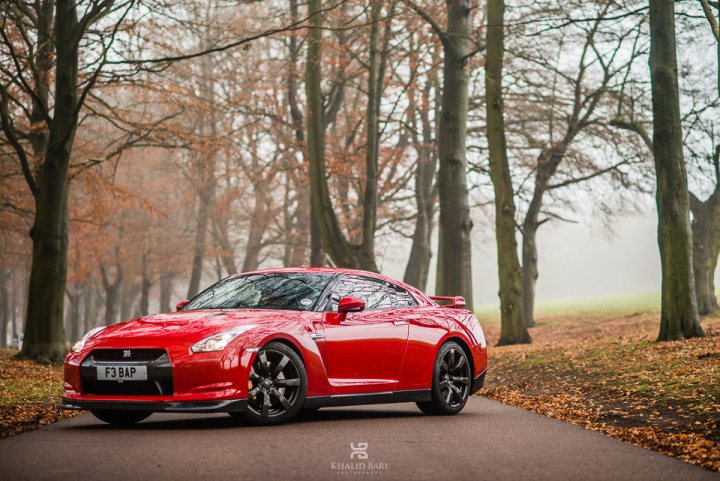 Some more stunning examples of Khalid Bari's work. - Page 1 - Herts, Beds, Bucks & Cambs - PistonHeads - The image captures a dynamic scene on a foggy road. A vibrant red sports car is the focal point, poised to move forward with its glossy finish reflecting the ambient light. The road stretches before the car, disappearing into a lush landscape of verdant trees and fallen leaves, scattered throughout the scene. The foggy environment enhances the mysterious and inviting atmosphere of the setting. A watermark reading "Touring" along with a series of numbers, 1467, is subtly visible in the background, indicating the photographer or creator of the image.