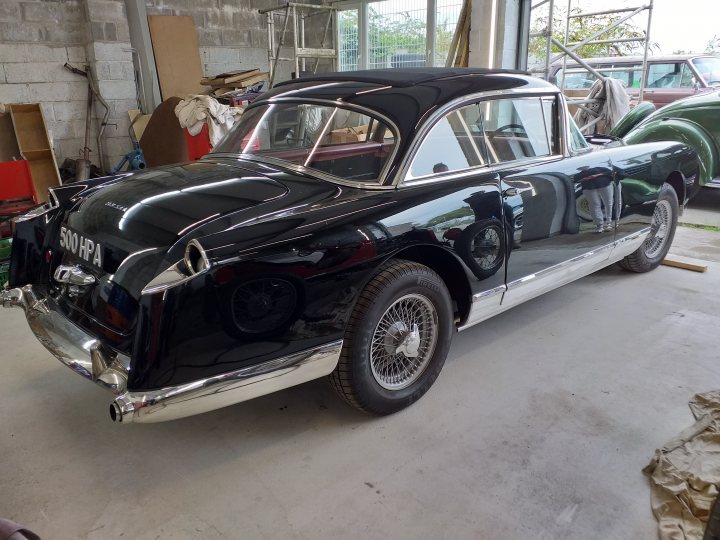 Webasto sunroof - fitting in 2021 to a classic - Page 3 - Classic Cars and Yesterday's Heroes - PistonHeads UK - The image shows a vintage car displayed indoors. The vehicle appears to be an older model, possibly from the mid-20th century, with a distinctive design featuring two doors and a prominent front grille. It has a classic black color scheme, which is consistent throughout the car's exterior. On the side of the car, there is a silver stripe running along its length. The car's wheels have a unique design with hubcaps. It is parked in front of another vehicle that appears to be an antique truck or van, suggesting this might be a display area for such classic vehicles.