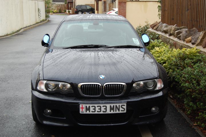 Lucky Luciano detailed my motor! - Page 1 - South West - PistonHeads - The image shows a black BMW car parked next to a wall, featuring rain-soaked car surfaces indicating recent rainfall. The car is oriented towards the viewer and is accompanied by a license plate stating "M33 MNS." To the right of the parked car, there is a row of orange rocks, partially obscured by the parked vehicle. The surroundings include a pavement and a fence, hinting at a residential or urban area. A portion of a person can be seen waiting to cross the street in the background, suggesting the photo was taken in a semi-busy area.