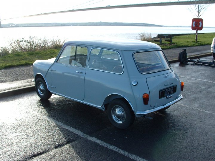 My 1960 Austin seven - Page 1 - Classic Minis - PistonHeads - The image features a scene of a small, blue car parked on the side of a road, which seems to have a rural or semi-urban setting. The vehicle is a compact model with a rounded design, and it appears to be in good condition, with no visible damage or scratches. It is parked facing the sea, with the Gentoo Islands visible in the distance across the water, which suggests that the car might be parked on a coastal road. The surrounding area includes a small patch of grass and a few trees, and there's a red sign mounted on a pole just behind the car. There's also a person in the background, though their involvement with the scene is not clear from this viewpoint. The overall lighting and reflection suggest it might be either early morning or late afternoon.