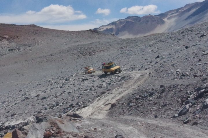 Pistonheads - The image shows a rugged terrain with a dirt path that has been partially cleared or bulldozed. A few yellow vehicles are on this path, possibly construction equipment or service vehicles. In the background, there is a rocky mountainous area covered in what appears to be snow or frost. The ground is uneven and rocky, indicative of a harsh environment. There's no visible vegetation, suggesting that it might be a high-altitude area or during a season when vegetation is scarce. The vehicles and the path seem to suggest some human activity in this remote location.