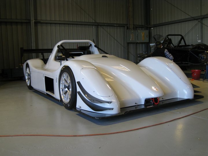 The image shows a white racing car parked indoors, likely in a garage or workshop. The car is sleek with a prominent aerodynamic nose and side pods that add to its streamlined appearance. It appears to be a single-seater vehicle, suggesting it may be used for professional racing or track-days. The car is either being prepared for a race or has just finished one, as evidenced by the open hood revealing the engine, though the engine itself is not clearly visible. There are no people visible in the image, focusing attention solely on the race car.