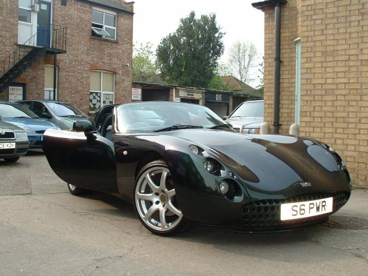 Your Best Tuscan Pic... - Page 1 - Tuscan - PistonHeads - The image shows a black sports car parked on a brick-paved street. The sports car is sleek, with a low, aerodynamic profile. It has a two-seater design and is parked on the right side of the street, facing left. There are other cars parked in the background, including a blue vehicle next to the building. A brick building with a visible staircase forms the backdrop of this scene.