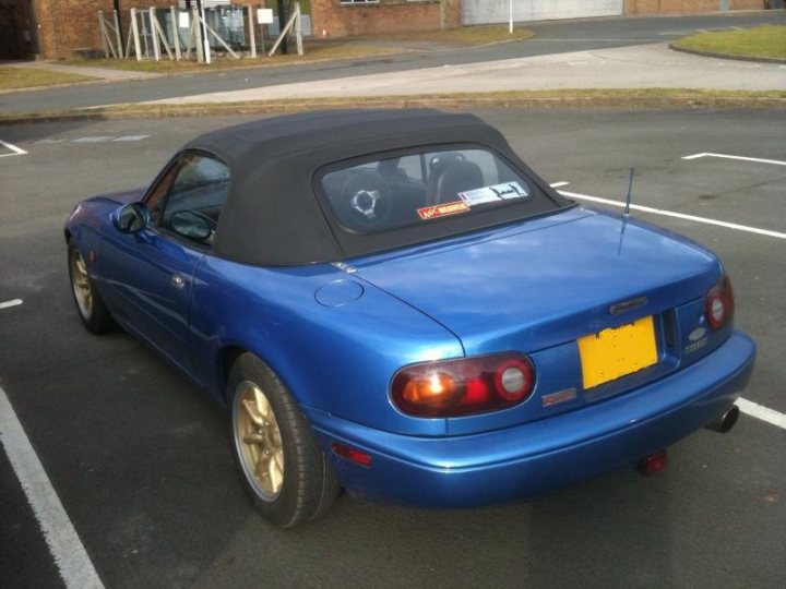 £3k, too steep? plus introduction - Page 1 - Mazda MX5/Eunos/Miata - PistonHeads - The image shows a blue convertible sports car parked in a lot paved with asphalt spots marked with white lines. The car is the central focus, with its metallic blue finish gleaming under the light. The top of the car is down, indicating the sun is out. There are several design factors that suggest performance, such as a rear spoiler and a rear wing. The license plate and a decal are on the back of the car, and there is a yellow plate near the license plate.