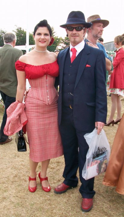 Pistonheads Revival Newbies - In the image, there is a man and a woman standing side by side on what appears to be grassy ground. The man is wearing a dark suit with a red tie, while the woman is dressed in a matching red dress with red shoes and a red handbag. Both individuals are smiling and posturing confidently. In the background, there are other people visible, some of whom are also wearing hats.