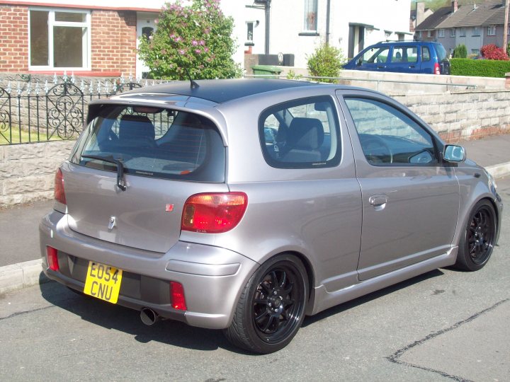 hi guys im new here...... - Page 1 - South Wales - PistonHeads - The image depicts a silver hatchback car with a license plate reading "E054 CNU" parked in a residential area. The car has black base rack, a spoiler, and dual exhaust pipes. To the right of the vehicle, there is a blue car parked on the same street. The scene is set alongside a row of houses, with a brick wall and a green bush nearby. There is also a side view mirror on the small car, indicating it is intended for one person to drive.