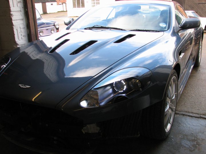 DB9 LED Lights - Page 1 - Aston Martin - PistonHeads - The image shows a black convertible sports car parked on a concrete surface with other vehicles nearby. The car's hood is up, revealing part of its engine. The background features a building and clear skies. There are no visible texts or distinguishing features in the image that provide additional context.