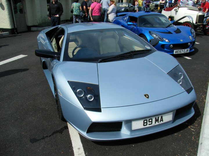 Breakfast Meeting, The Hare, Roxwell 4th June - Page 1 - Kent & Essex - PistonHeads - This image showcases a metallic blue sports car parked on a black asphalt surface with white lines marked on the road. The car is positioned at an angle, with its side facing the camera, revealing shiny reflective surfaces and the chassis of the vehicle. Another sports car is faintly visible in the background to the right, and a group of people can be seen behind the blue car, mingling and observing the cars. The setting appears to be a public parking area or an open road where motorists can park and possibly engage in conversation.