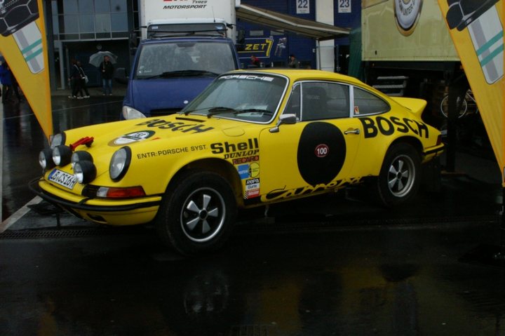 Pistonheads - The image captures a vibrant yellow Porsche 911 GT1 exemplum, proudly showcased with the words "Bossco Shell" emblazoned at the front and a striking black and white circular pattern on the side. This vehicle is specifically designated as an "Enter Porsche System," suggesting it might be part of a racing team or showcase event. The Porsche is prominently displayed within a clearly marked parking area, a direct contrast to the urban setting visible in the background with buildings and other vehicles. A number of people and umbrellas are present, indicating that this might be a public event or a special occasion where the car is on display.