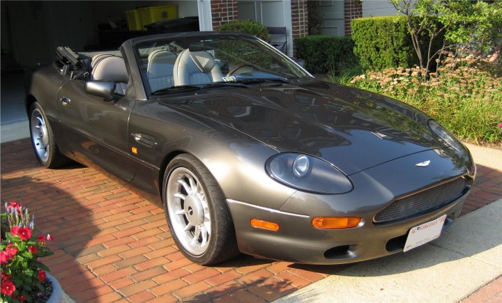 Girl Fancy Loaded Pistonheads Single - The image shows a parked sports car with a convertible top, suggesting it's an open-top model, likely from the Jaguar or similar brand. The car's body is dark in color, possibly black or dark gray. It's positioned on a brick driveway, which adds a classic, red-brick paved surface to the scene. To the left of the car, a small garden is visible with plants and what appears to be a bird feeder. The sun is shining, casting shadows and highlighting the glossy finish of the car. The setting suggests a residential area, possibly during the early morning or late afternoon, given the low sunlight level.