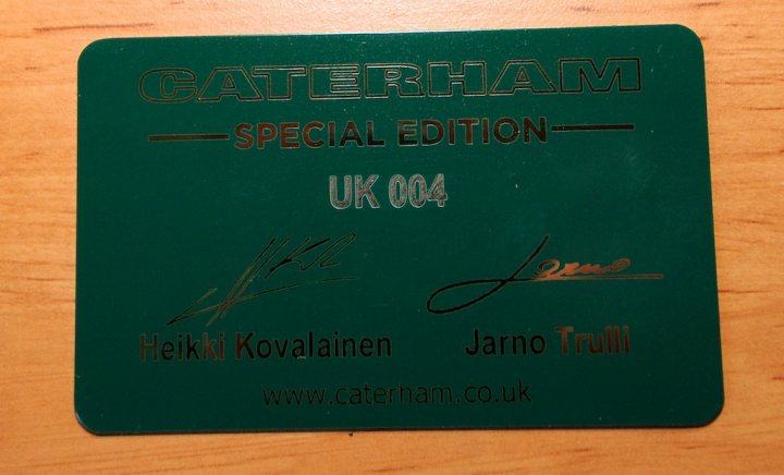 My R500 Team Lotus Special Edition - Certificate and Plaque - Page 1 - Caterham - PistonHeads - The image shows a green, rectangular card with metallic details on a wooden surface. The card reads "Caterham Special Edition UK04" in a shiny, gold-colored font on the upper part, leaving ample space below. The lower part features the names "Heikki Kovalainen" and "Jarno Trulli" in cursive, black lettering. The bottom of the card also displays a website address: "www.caterham.co.uk." The style of the card suggests it might be a collector's item or a commemorative piece related to racing or automotive history.