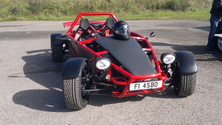 Help identifying kit car - Page 1 - Kit Cars - PistonHeads - The image depicts a red, three-wheeled ATV parked outdoors. The vehicle is small with a compact wheelbase, and it features large, over-sized wheels that contrast with its bright red body. Above the windshield of the ATV, there are four backrest seats, suggesting it's a single-seater vehicle designed for multiple riders. The ATV is standing on a surface that appears to be a parking lot or similar concrete area, with patches of grass visible in the periphery.