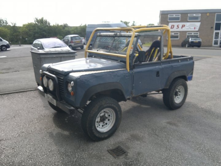 Pistonheads - The image shows a vintage blue off-roader truck parked in a lot. The truck has a yellow spare tire rack on top, suggesting it may be used for transporting sporting equipment. A building with the letters "DSM" visible can be seen in the background, indicating this might be a commercial or industrial area. The truck has a distinguished, robust appearance, typical of such vehicles from a bygone era. It faces towards the viewer, creating a clear view of its front. The ground shows visible tire marks and has some dark stains, possibly from gas or oil, suggesting the truck may have been parked there for some time.