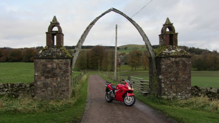 VFR as a first bike? - Page 2 - Biker Banter - PistonHeads - The image features a vibrant red motorcycle parked on a narrow dirt road. The motorcycle, noticeable with its headlights on, is positioned on the side of the road, directly under a worn-out stone archway. The archway is identical on both ends and is adorned with similar architecture and stone details. Beyond the archway and the motorcycle, a lush green field can be seen, suggesting a rural or countryside setting. The sky above is slightly cloudy, indicating that the photo was taken on an overcast day.