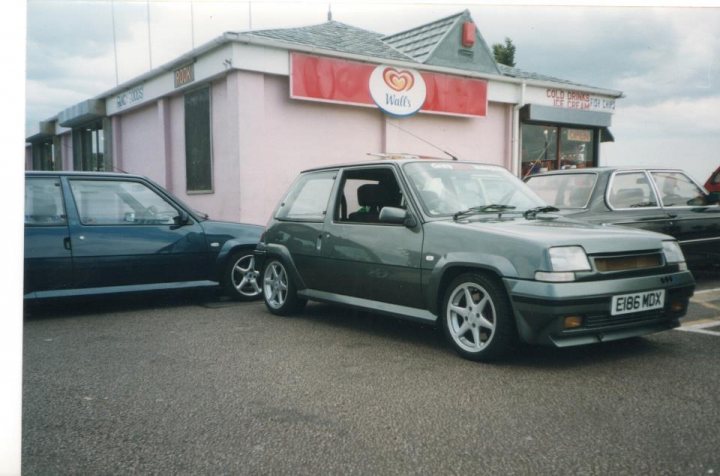 R5 GT Turbo  - Page 2 - French Bred - PistonHeads - This image depicts a gray hatchback car parked in front of a building. The car is positioned fairly close to a black car in the adjacent space. On the building, there is a large red sign with a heart-shaped white logo and white text that reads "WICK'S". In the background, the sky is partly cloudy, suggesting a somewhat overcast day. The cars and the sign convey a sense of a commercial or retail area. There are also small figures that appear to be pedestrians in the far background of the image.