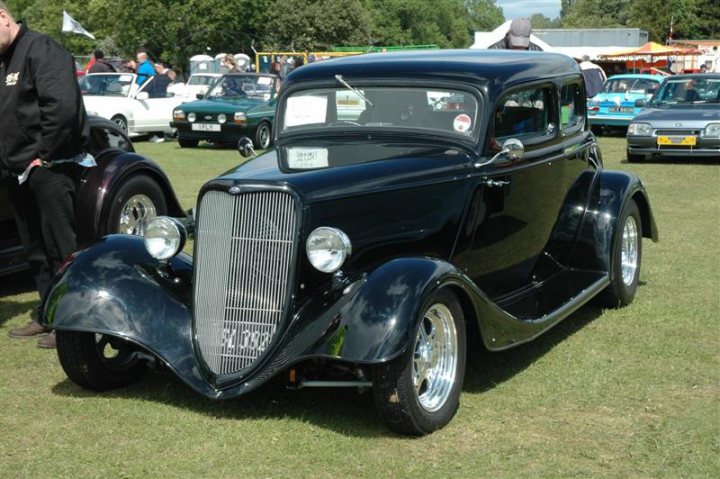 Fins and Chrome - Page 1 - North East - PistonHeads - The image showcases a nostalgic car show where a variety of classic cars are on display. The focal point of the image is a vintage black-and-white sedan, distinct for its shiny grill and imposing chrome bumper. This sedan is parked amidst other classic cars, suggesting a gathering of automobile enthusiasts sharing their passion for vintage vehicles. In the background, people can be seen admiring the showcars, their presence adding a lively atmosphere to the event.