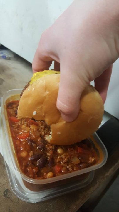 Photo of your dinner (vol 2) - Page 455 - Food, Drink & Restaurants - PistonHeads - In the image, a person's hand is seen grabbing a sandwich that's placed on top of a container full of chili. The sandwich appears to be the centerpiece of the image, prominently displayed on the bowl. The bowl itself is set on a table, and below it, there's a stack of plastic containers, possibly for storage or serving. The background is largely out of focus, keeping the focus on the hand and the sandwich.