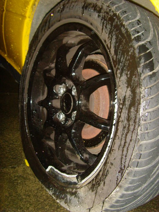 Wheel Refurbrepair Pistonheads - The image shows a close-up view of a vehicle's wheel and tire. The tire appears to be an older model, with visible signs of wear, including a tread pattern that suggests it has been used for some time. There is a tan surface reflecting the wheel, indicating the photo might have been taken on a dirty or muddy ground. The wheel is black and steeped in mud, which extends into the gaps and edges of the wheel itself. There is no visible text or branding in the image. The style of the image is a short, targeted photograph focused on the wheel and tire.