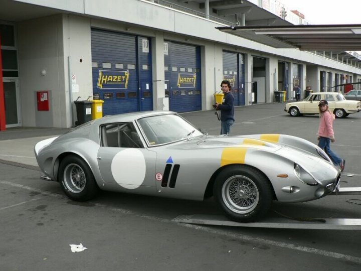 Silver, yellow stripe - Page 1 - Elise/Exige/Europa/340R - PistonHeads - The image showcases a striking daytime scene where a gray and white sports car is parked, with the hood raised, in front of a garage facility. The car is sleek and stylish, bearing the number 1 on its side, indicating it might be a racing vehicle or part of a racing team. A couple of individuals are standing nearby, likely enjoying a quiet moment on this sunny day. The background reveals rows of cars parked and additional garage structures, creating a sense of depth and perspective.