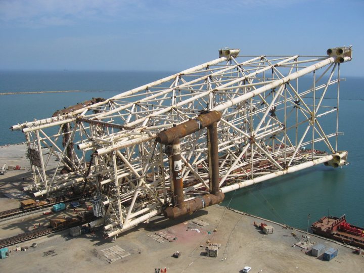 Post pictures of amazingly cool engineering - Page 21 - Boats, Planes & Trains - PistonHeads - The image shows a large, complex white structure with robust towers and an intricate network of horizontal bracing and vertical supports. It appears to be a crane barge on dry land, as indicated by the presence of water and a ship in the background. The perspective suggests the photo is taken from a bird's-eye view, providing a comprehensive view of the structure's size and design. The crane barge, possibly used for offshore drilling or construction, seems dismantled or partially disassembled, given the absence of superstructures or cabins typically seen on such equipment when operational.