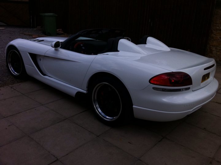 Those side exit exhausts... - Page 1 - Vipers - PistonHeads - The image shows a white sports car parked on a brick driveway. The car is a convertible, ready for the top down experience. From the angle of the photo, the car appears pristine and shiny, suggesting it's well-maintained. The background is minimal, featuring what looks like a garage door, which gives context to the location of the vehicle. The lighting differs across the image, with darker areas that contrast with the brightness on the car's surface, creating an interesting interplay of light and shadow.