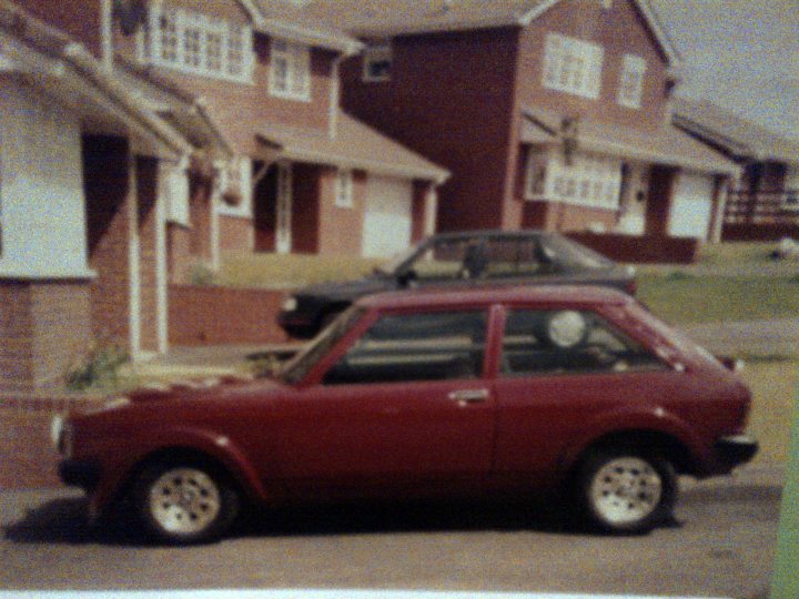 A car parked in front of a building - Pistonheads - The image portrays a serene suburban neighborhood. A vibrant red compact car is parked on the street, facing towards the right. The car's shiny exterior reflects the ambient light, highlighting its sleek design. The street where the car is parked is lined with houses, their red brick facades reinforcing a sense of uniformity. The image has a vintage filter applied, giving it a warm and nostalgic feel.