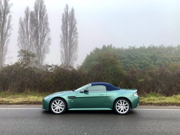 The non-black-non-grey-non-silver thread - Page 46 - Aston Martin - PistonHeads UK - The image shows a blue convertible sports car on a road. The vehicle is parked, facing the viewer with the top down. It has white-walled rims and is equipped with a sunroof, which is partially open. The car's design includes a sleek body and a prominent front grille. The background features a misty or foggy landscape with greenery and a hint of a building. There is no visible text on the image.