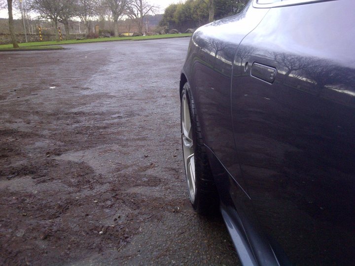 DBS wheels on DB9 - (more or less) - Page 1 - Aston Martin - PistonHeads - The image shows a close-up view of a black car. The car's hood is captured in the foreground. The car appears to be parked on a lot with a muddy surface or a large puddle. In the background, there is a field with trees and a park-like setting. The photo is taken on a cloudy day.