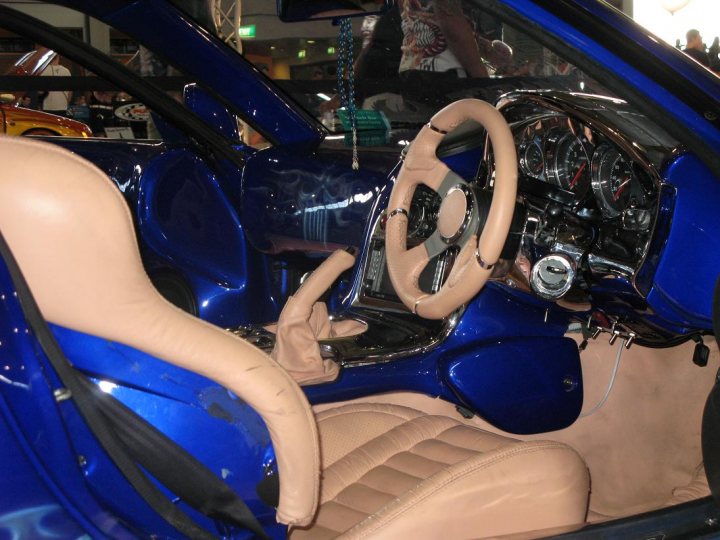 Pistonheads Autosalon Sydney - The image features the interior of a blue sports car, exhibited in a showroom. The focus is on the driver's side of the dashboard, where the steering wheel and column are visible. The sporty seats are upholstered in a beige leather, providing a contrast to the blue exterior. In the mirror, there's a reflection of the outside light, and the gear shift is prominently located on the side, ready for engagement. Another car is visible in the background, located beyond the showcased sports car.