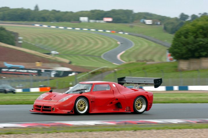 help with cooling - Page 1 - Ultima - PistonHeads - The image captures a dynamic scene on a racetrack. Dominating the frame is a red race car, its aerodynamic shape designed for high speeds. The car is moving along a winding track, its speed evident in the slight blur of the tires. The racetrack itself is situated in a valley, surrounded by lush green hills, providing a stark contrast to the mechanical beasts intent on scorching them. Above, the sky is a clear blue, suggesting a beautiful day for racing. In the distance, the silhouette of the grandstand and other racing circuits can be seen, adding depth and context to the scene.