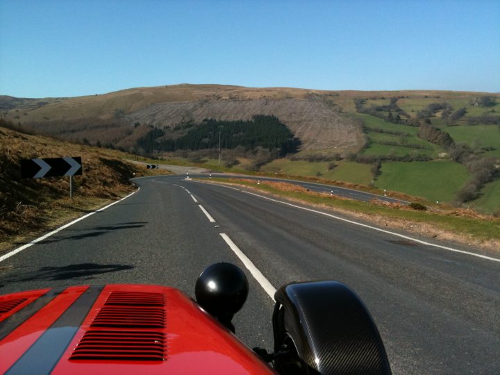 Wales Blat Pistonheads - The image captures a picturesque scene with a nearly empty two-lane road. There is a red vehicle on the right side of the road, facing the camera. The backdrop consists of a lush green hill or hillside lined with trees, providing a serene rural landscape. Above, the sky is a clear blue, and there's a solitary triangle sign on the left side of the road. The overall atmosphere of the image is tranquil and open.