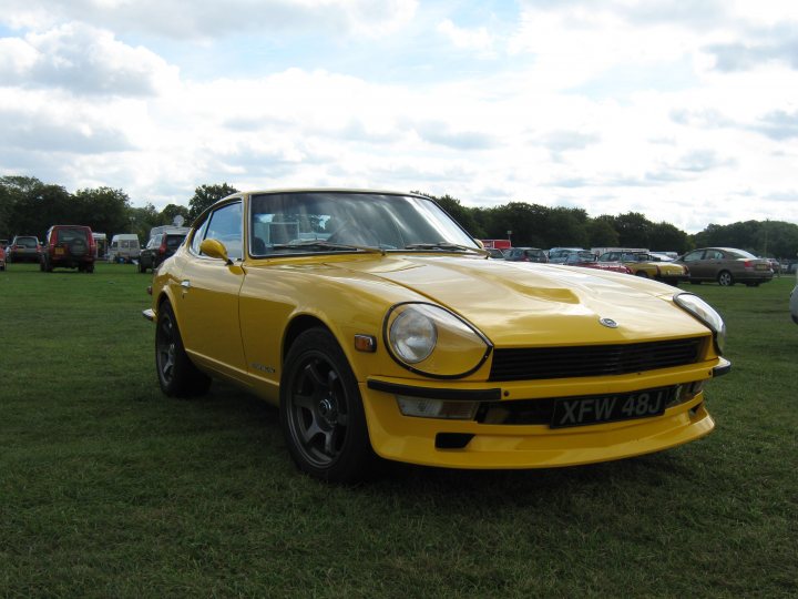 Datsun 240 or 260Z - Page 1 - Classic Cars and Yesterday's Heroes - PistonHeads - The image showcases a vibrant yellow Datsun 240Z sports car parked on a grassy field. The vehicle's sleek design, characterized by its curved rear edges, is accentuated by the black and white license plate. The Datsun 240Z's silver emblem and dual headlights are also noteworthy. The car is the main subject in the photograph, captured in broad daylight. The background reveals a community event, with various other cars parked in a parking lot, suggesting the Datsun 240Z is part of a larger gathering of automobile enthusiasts.