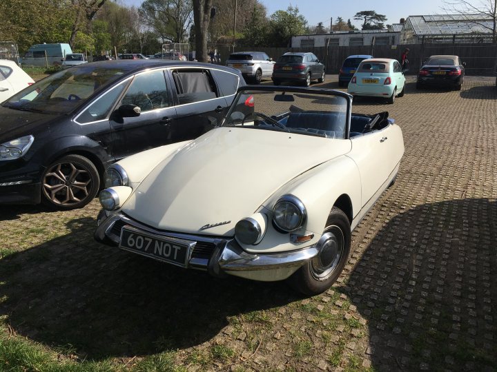 The BAD PARKING thread [vol3] - Page 463 - General Gassing - PistonHeads - In the image, a white BMW convertible car is parked on a gravel driveway on a sunny day. The car's top is down, revealing a red interior. In the background, there are several other parked vehicles, including a black car and a turquoise car. Additionally, there is a person standing towards the right side of the image. A portion of a building can also be seen in the far background.