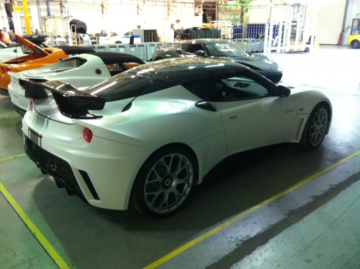 I got one! - Page 2 - Evora - PistonHeads - The image is a photograph of a white supercar parked inside a garage or an indoor storage space. The vehicle is sleek with aerodynamic bodywork and features large side intakes and a prominent rear wing. It is fitted with five-spoke wheels, and there is a green and yellow stripe visible on the floor around the car. The setting is fairly industrial, with a backdrop of other cars and equipment. The lighting within the space is artificial, casting shadows that contribute to the overall mood of the image.