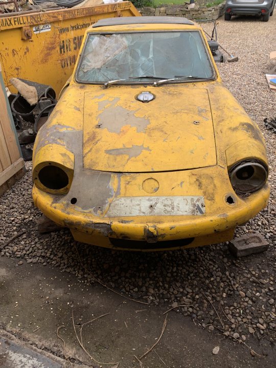 Classics left to die/rotting pics - Vol 2 - Page 340 - Classic Cars and Yesterday's Heroes - PistonHeads UK - The image shows a classic sports car parked outside. It appears to be a yellow convertible with a hard top, featuring an unusual front-end design. The car is situated on a driveway next to a building, and there are several other cars visible in the background. The vehicle's condition suggests it might be a project or restoration, as indicated by its worn appearance and missing parts such as the hood. The setting seems to be a private property or a repair workshop given the presence of what looks like industrial or construction equipment.