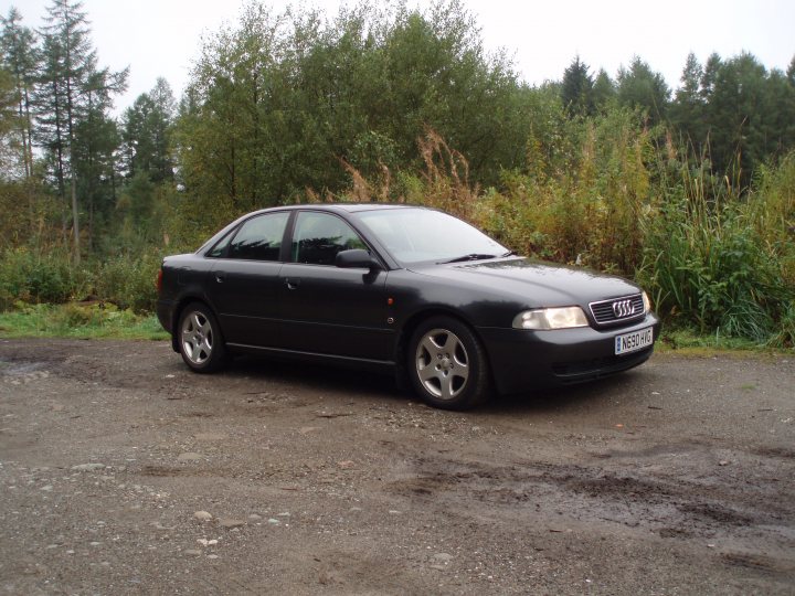 My old Audi A4 Sport 1.8T 20v 150bhp - The image shows an Audi car parked on a gravel surface. The car is predominantly black, and its left side is visible. The setting appears to be a wooded or natural area as there are trees and vegetation in the background. The sky is overcast, suggesting an overcast day. The ground is covered with small rocks. The car's design and the surrounding forest give the image a peaceful, serene atmosphere.