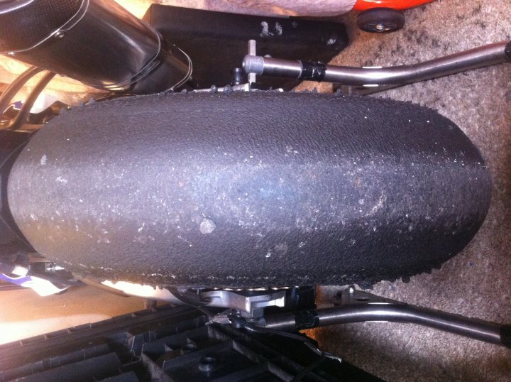 the bb trackday thread.   - Page 11 - Biker Banter - PistonHeads - The image presents a black tire with a visible tread pattern, placed in a room with a carpeted floor. The tire has a few scratches and spots, suggesting it has been used. The tire is separated from its usual positioning, such as on a car or motorbike, and is in close-up view against a black background. The setting appears to be an indoor environment, with mechanical parts and a metal structure hinting at a maintenance or manufacturing setting.