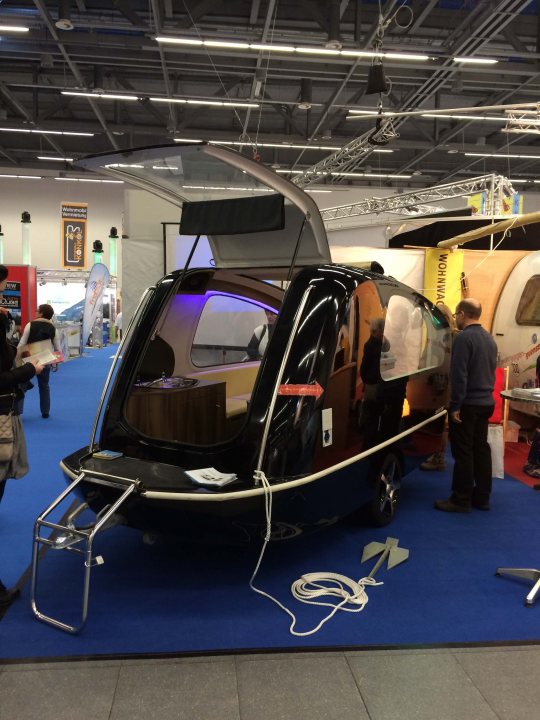 A group of people standing next to a train - Pistonheads - The image displays an indoor setting, possibly a trade show or exhibition, where a small, futuristic-looking hovercraft or air car is the central attraction. Surrounding the vehicle, several people are seen, some standing close to the daring piece of equipment while others observe from a distance. There are also small vehicles or machines that might be related to the hovercraft or part of the display. One man is standing next to the hovercraft, engaged in conversation with a woman who is facing away from the camera. The weather inside is dimly lit, giving the overall atmosphere an ambient, modern vibe.