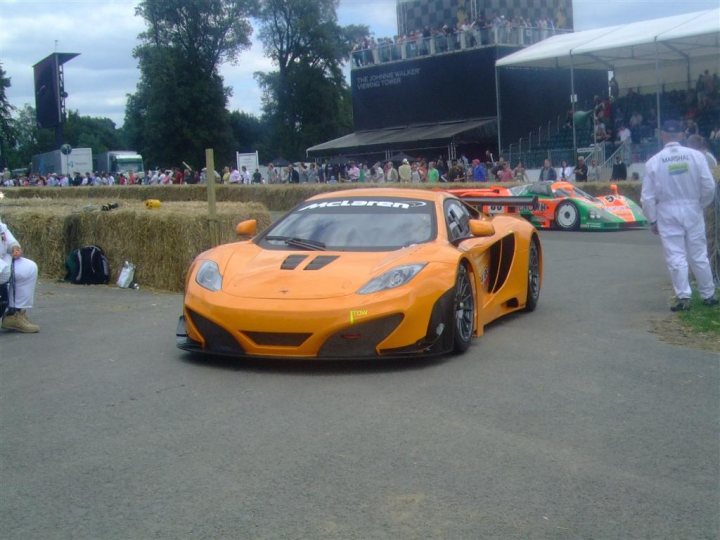 Goodwood FoS pics / videos - Page 1 - Goodwood Events - PistonHeads - The image presents a vibrant scene from a race track. The focal point is an orange racing car, centered in the frame, with its hood slightly raised, revealing engine monitors. This car is poised next to another green vehicle, their sleek designs indicative of high-performance engineering. The track is encased by towering trees and an audience can be seen in the background, creating a dynamic backdrop to the image. The atmosphere is intense and exciting, capturing the essence of a racing event.