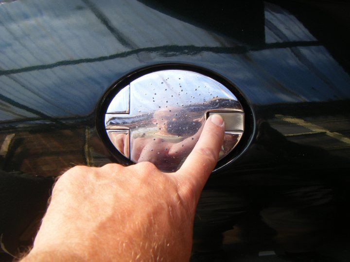 Fuel Filler Cap Security Idea - Page 1 - S Series - PistonHeads - The image captures a moment where a human hand is visible, pressing down on a car door handle. The handle is silver, and it's reflecting in the small circular window on the car door, possibly a window lock mechanism. In this reflection, the bottom of a human face is visible. The car door is black and appears to be made of a shiny material, which suggests it might be a car that's parked in a way that the door is facing the viewer. The overall setting seems to be an outdoor scene with some natural light reflecting on the car's surface.