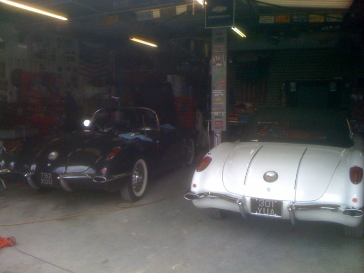 Services Pistonheads Ian Goss - The image depicts a spacious garage with a high ceiling and multiple bays. Two vintage cars are the main subjects, parked side by side near the middle of the frame. The car on the left, with its pointed nose and weathered appearance, could be an early model, possibly from the 1950s or 1960s. The car on the right has a sleeker design, possibly from the 1970s or 1980s, and both vehicles have a touch of color contrasting with their predominantly black and white exteriors. The garage is well-lit, with natural light filtering in from a large window or skylight, and the overall scene suggests a place where restoration or maintenance might take place for classic and specialty vehicles.