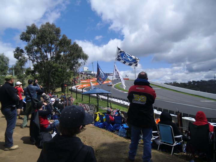 Bathurst 1000 - Page 1 - Australia - PistonHeads - The image captures a vibrant scene at a race track. Numerous spectators are spread around the course, their faces turned towards the track where a race is in progress. The spectators are closely packed together, with some sitting on chairs while others stand on the grassy hillside. The sky overhead is cloudy and blue, suggesting it might be a cool day. In the background, large flags from different racing teams add a festive atmosphere to the event.