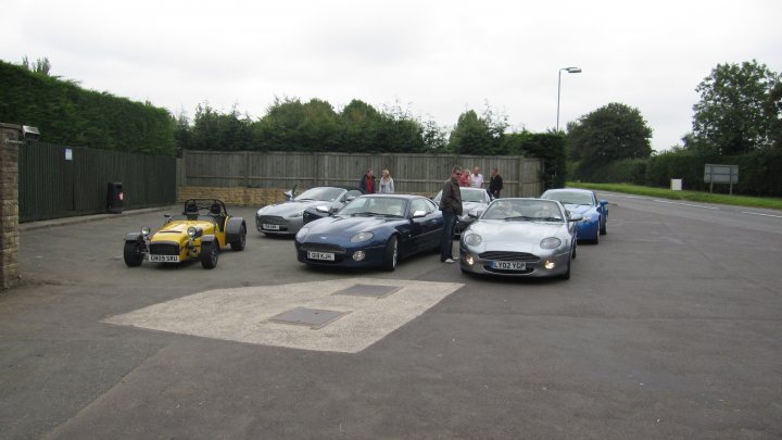 Pistonheads - The image depicts a line of cars parked in an open area, possibly a parking lot. The cars are of different models and colors. Behind the vehicles, multiple people are seen. To the extreme left, an orange vehicle is visible with a person standing near it. The ground appears to be made of concrete or asphalt, and there are hedges that line the longest side of the area.