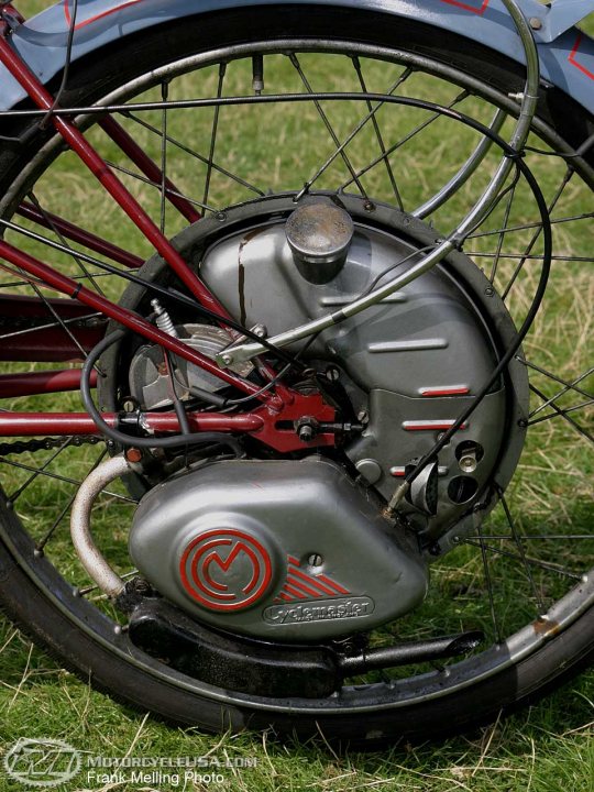 Petrol powered mountain bike legallity? - Page 1 - Speed, Plod & the Law - PistonHeads - The image shows a close-up view of the chain mechanism on a bicycle. The bike's wheel and frame are partially visible. The chain is secured to a sprocket, which is attached to a gearbox. The photo has a watermark that reads "MotorcycleUS.com" and "Frank Melling," indicating that the image is likely from a website specializing in motorcycles. The focus is on the rear wheel and sprocket, captured against a backdrop of grass, emphasizing the mechanical nature of the scene.